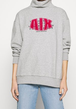Grauer Pullover mit Stehkragen, gekennzeichnet durch ein auffälliges pinkes "AIX"-Logo und schwarzen Text. Lockere Passform mit gerippten Bündchen und Saum. Kombiniert mit hellblauen Jeans.
