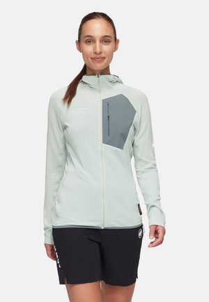 Veste zippée vert clair avec un tissu texturé, dotée d'une poche poitrine grise et d'une capuche. Associée à un short noir affichant des logos de marque.