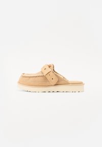 Chaussure slip-on en suede beige avec des coutures de style mocassin, sangle ajustable et semelle en caoutchouc blanche épaisse, présentée sur un fond blanc.