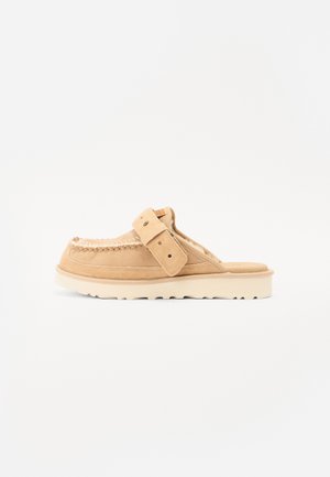 Beige suede slip-on shoe with moccasin-style stitching, adjustable strap, and thick white rubber sole, shown against a white background.