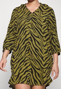 Sukienka z krótkim rękawem w oliwkowozielonym kolorze z czarnymi paskami tygrysa. Posiada dekolt w serek oraz luźny krój z zaokrąglonym hemem.