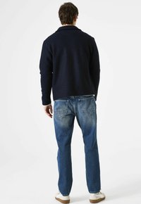 Man in een marineblauwe jas, met blauwe jeans en witte sneakers, staand met zijn rug naar de camera tegen een effen witte achtergrond.