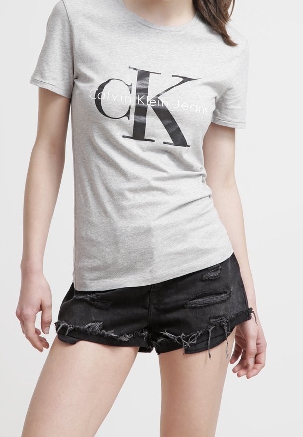 Žena oblečená v šedém tričku Calvin Klein Jeans a černých roztrhaných denimových šortkách na jednobarevném pozadí.