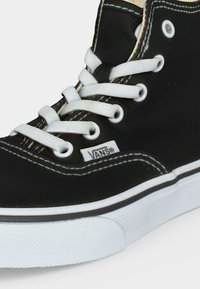 Μαύρα ψηλά αθλητικά Vans με λευκά κορδόνια, λευκή ραφή και λευκή λαστιχένια σόλα σε απλό φωτεινό φόντο.