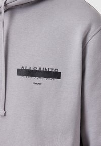 Grijze hoodie met een relaxte pasvorm, verstelbare trekkoordkap en duidelijke grafische tekst in het zwart, inclusief merknaam en locatie.