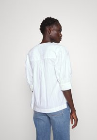 Hvit, stripete bluse med pufferminner, rund hals og plisseringer bak; har oppbrettede ermer med knappdetaljer.