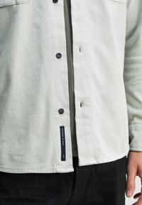 Close-up van een lichtgroen knoopshirt gedragen over een bijpassend T-shirt, gecombineerd met zwarte broek, met een "Butcher of Blue" label.