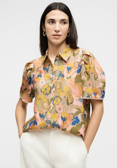 Γυναίκα φορόντας κοντομάνικο φλοράλ μπλουζάκι με φουσκωτούς ώμους και λευκό παντελόνι, στέκεται με τα χέρια στις τσέπες μπροστά σε απλό φόντο.