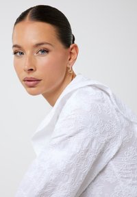 Blusa bianca ricamata con motivi floreali, colletto alto e tessuto strutturato. Orecchini a cerchio dorati completano il look. Design pulito e moderno.