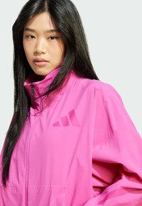 La chaqueta rosa con cremallera presenta un alto cuello, grandes bolsillos delanteros y un logo en relieve en el pecho. Tejido ligero con una textura suave.