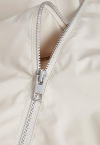 Beige zip-up jakke med en sølvglinsende glidelås og glatt stoffoverflate, med et nærbilde som fremhever glidelåsens detaljer og sømmer.