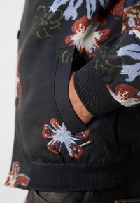 Χέρι στην τσέπη ενός σκούρου ζακέτας με floral σχέδιο με ριγέ μανσέτες και μέση, που διαθέτει μεγάλες κόκκινες και μπλε άνθη.