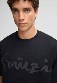 Schwarzes Baumwoll-T-Shirt mit glänzendem schwarzen Logodesign auf der Vorderseite. Klassischer Rundhalsausschnitt mit kurzen Ärmeln. Glatte Textur.
