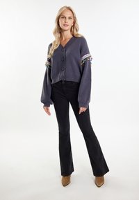 Cardigan grigio a maglia con scollatura a V, cinque bottoni e bordo colorato con frange sulle maniche. Abbinato a pantaloni neri svasati e stivaletti color cammello.