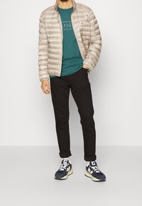 Beige puffatakki, teal-T-paita graafikolla, mustat housut ja tummansinisiä lenkkareita, joissa on valkoisia yksityiskohtia. Sileät pinnat kauttaaltaan.