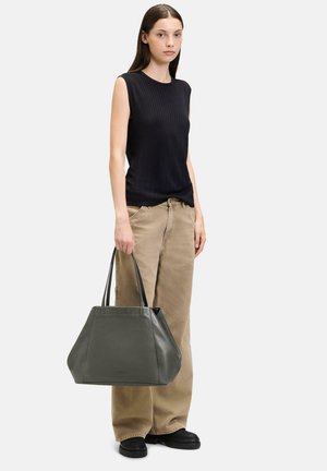 Gray leather tote bag with black handles, angular shape, and visible stitching. Model wears a black ribbed top and beige trousers.