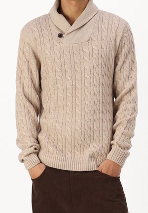 Beiger Strickpullover mit Zopfmuster, Schalkragen und einem Knopf, getragen mit den Händen in den Taschen einer dunkelbraunen Cordhose.