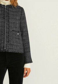 Schwarze und weiße Tweedjacke mit cropped Design, strukturierter Stoff, Reißverschluss vorne und fransigen Kanten, ausgestattet mit Seitentaschen und langen Ärmeln.