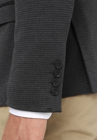 Rękaw teksturowanego, ciemnoszarego żakietu z czterema czarnymi guzikami. Widoczny mankiet białej koszuli poniżej. Dopasowany krój, strukturalny design.