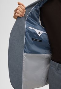 Grijs patroonblazer met een blauwe satijnen voering, voorzien van een wit biesje op de zak en een label met de tekst "FINEST STRETCH FABRIC."