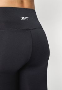 Czarne legginsy z wysokim stanem, wykonane z gładkiego, elastycznego materiału, z białym logo na pasku i płaskimi szwami.