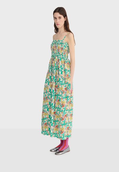 Vrouw met lang bruin haar, gekleed in een mouwloze bloemen maxi-jurk, roze gestreepte sokken en zilverkleurige metalen platte schoenen, staand opzij.