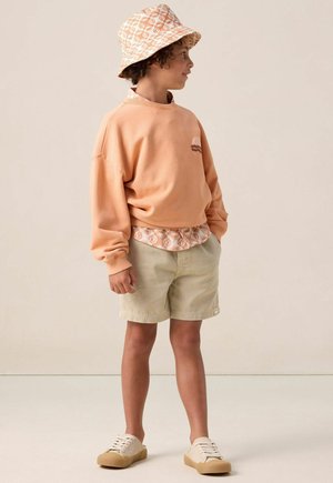 Oranges Sweatshirt mit einem kleinen braunen Grafik, grüne Shorts und einen gemusterten Bucket Hat. Die Schuhe sind beige Leinenschuhe mit einer strukturierten Sohle.
