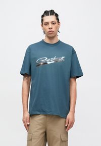 Camiseta de algodón color teal con mangas cortas, que presenta un logo metálico plateado "Carhartt" en el pecho. Diseño sencillo y cuello redondo.