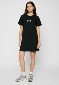 Robe t-shirt en coton noir avec des manches courtes, présentant un design de texte blanc "prier". L'ourlet atteint le milieu de la cuisse. Portée avec des baskets grises et des chaussettes blanches jusqu'aux chevilles.