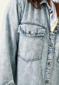 Ljust blå denimskjorta med två bröstfickor, silverknappar och en sliten yta. Har kontrasterande sömmar och en mönstrad etikett.