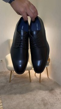 Main tenant une paire de chaussures habillées en cuir noir brillant avec des lacets, positionnées au-dessus d'un tapis beige et d'une chaise blanche.