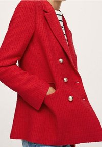 Veste rouge croisée à double boutonnage avec tissu texturé, boutons argentés et poches latérales. Un revers cranté complète sa silhouette cintrée.