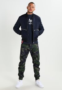 Veste bombardier navy avec des fermetures éclair argentées, associée à un pantalon cargo camouflage vert. Le mannequin porte des baskets blanches et un haut noir en dessous.