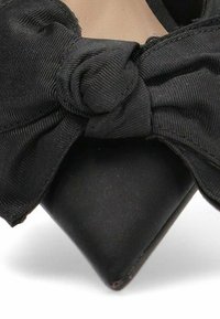 Zapato de tacón alto negro con un lazo texturizado y un nudo retorcido. El tacón es delgado y puntiagudo, hecho de un material liso y brillante.