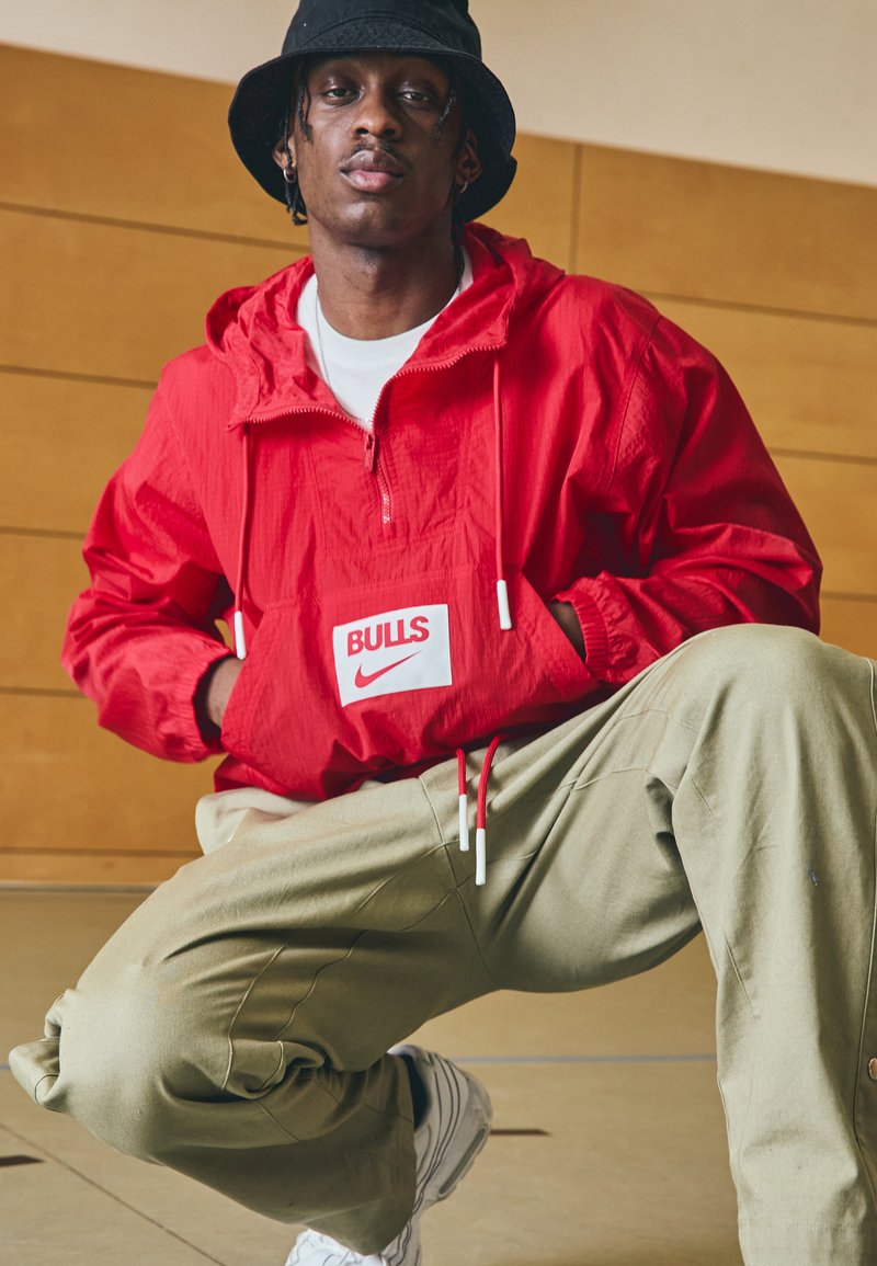 Röd Nike vindtät jacka med dragkedja vid halsen, "BULLS"-logotyp och vita accenter. Kombinerad med ljusgröna byxor och vita sneakers.