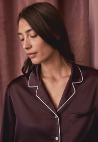 Burgundowa satynowa góra piżamy z białymi lamówkami, z kołnierzykiem z wycięciem, z przodem zapinanym na guziki oraz kieszenią na lewym piersi. Gładka tekstura.