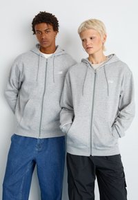 Felpe grigie con zip e cappuccio, dotate di cordini, tasche frontali e un logo. I modelli indossano jeans a contrasto e pantaloni cargo scuri.