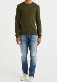 Olivově zelený pletený svetr s kulatým výstřihem, spárovaný s světle modrými vybledlými džíny a bílými teniskami. Jednoduchý design s žebrovanými manžetami.