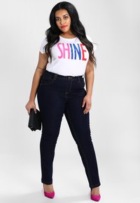 Bijela majica s natpisom "SHINE" u ružičastoj i plavoj boji, uparena s tamnoplavim uskim trapericama i ružičastim visokim potpeticama; drži crnu clutch torbicu.