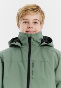 Grüne Sportjacke mit hohem Kragen und Reißverschluss, mit schwarzem Akzent auf der Vorderseite und Logoplatzierung auf der linken Seite. Einfaches Design.