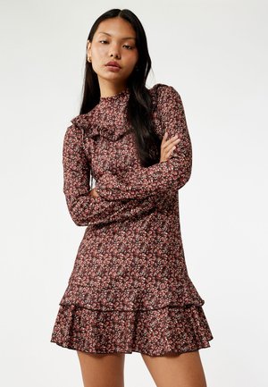 Bloemjurk met lange mouwen; heeft een hoge gerimpelde kraag en een volant aan de zoom; zwarte en diep rode basis met kleine roze en witte bloemen.