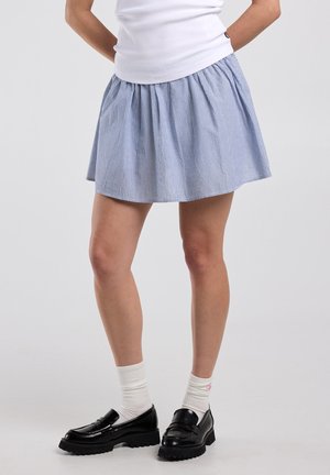 Beine in einem blau-weiß gestreiften Rock, weißen Socken und schwarzen klobigen Loafers, stehend vor einem einfachen hellen Hintergrund.