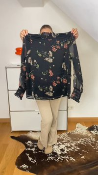 Blusa preta transparente com padrões florais em tons de rosa e verde. Apresenta mangas longas e bufantes e um decote redondo. Tecido leve.