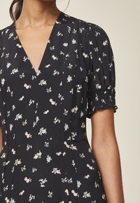 Svart klänning med djup V-ringning, korta puffärm och spridd blommönster i pastellfärger. Slät textur, figurnära silhuett.