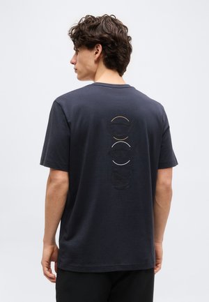 Nooruk mees tume lokkis juustega, kes kannab musta T-särki, millel on tagaküljel kolm reljeefset kohvitassi kujundit ja poolringid.