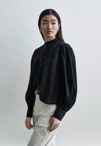 Γυναίκα με μακριά σκούρα μαλλιά που φοράει κρίκους στα αυτιά, μια άνετη μαύρη μπλούζα με φουσκωτά μανίκια και ανοιχτόχρωμο παντελόνι με ψηλή μέση, κοιτάζοντας πλάγια.