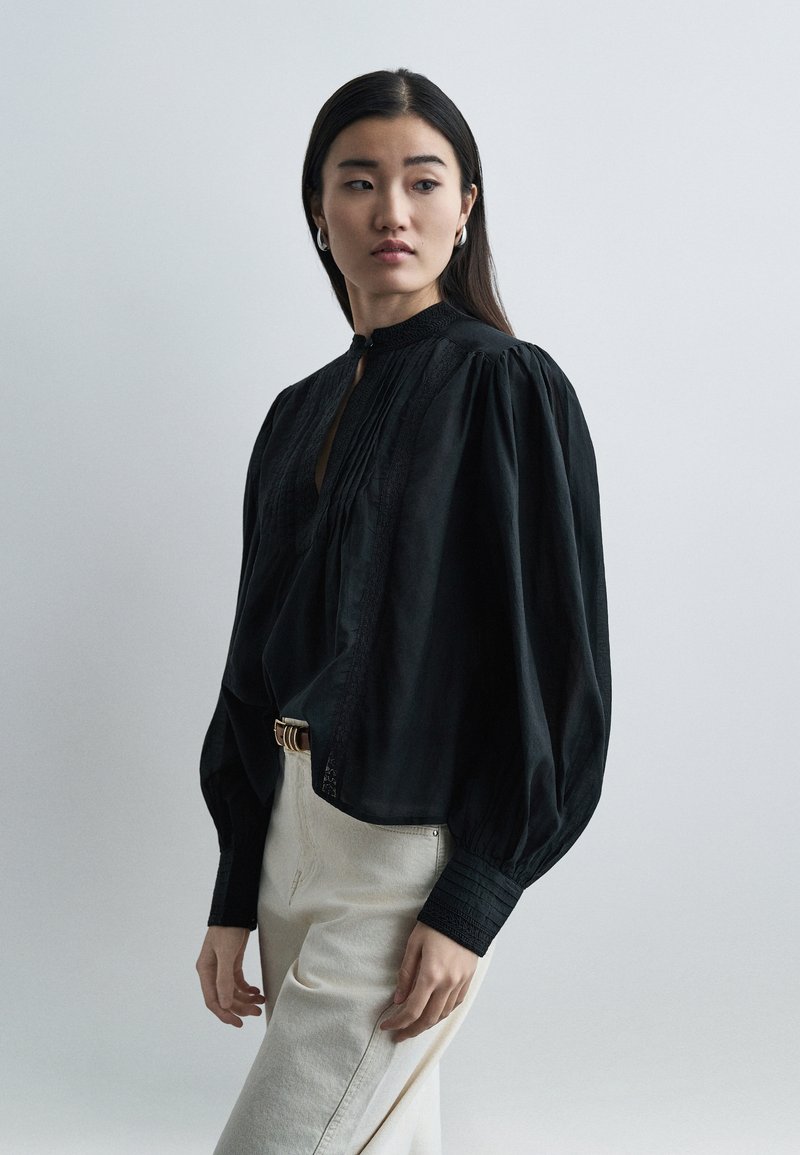 Γυναίκα με μακριά σκούρα μαλλιά που φοράει κρίκους στα αυτιά, μια άνετη μαύρη μπλούζα με φουσκωτά μανίκια και ανοιχτόχρωμο παντελόνι με ψηλή μέση, κοιτάζοντας πλάγια.