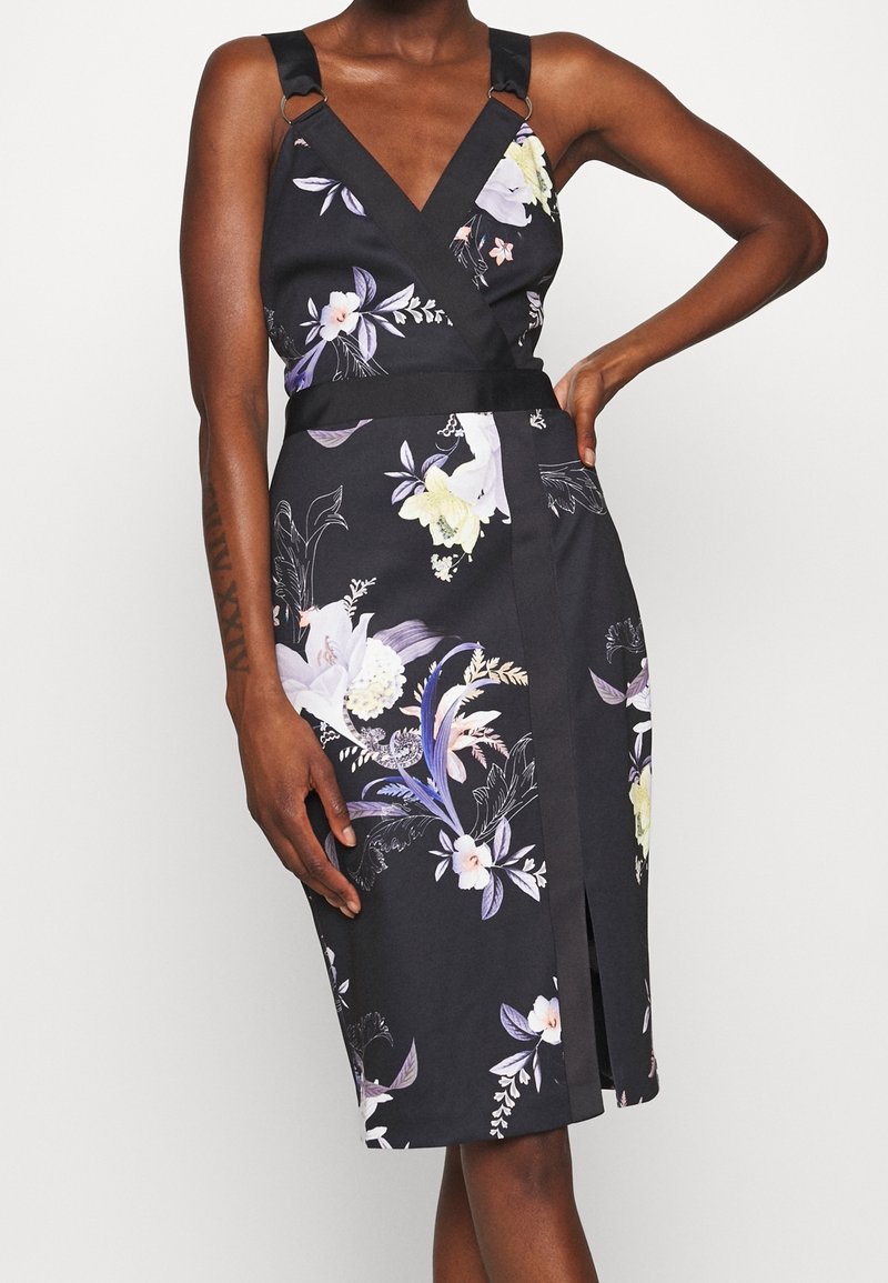 Femme portant une robe portefeuille noire sans manches à fleurs, avec de larges bretelles, un décolleté en V et une fente avant arrivant au genou.