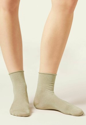 Paire de jambes portant des chaussettes courtes beige clair, debout sur un fond neutre.