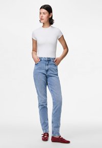 Λευκό εφαρμοστό μπλουζάκι, ανοιχτό μπλε τζιν με ίσια καλύμματα και κόκκινα παπούτσια με κορδόνια. Το μοντέλο ποζάρει με τα χέρια στις τσέπες του τζιν μπροστά σε απλό φόντο.